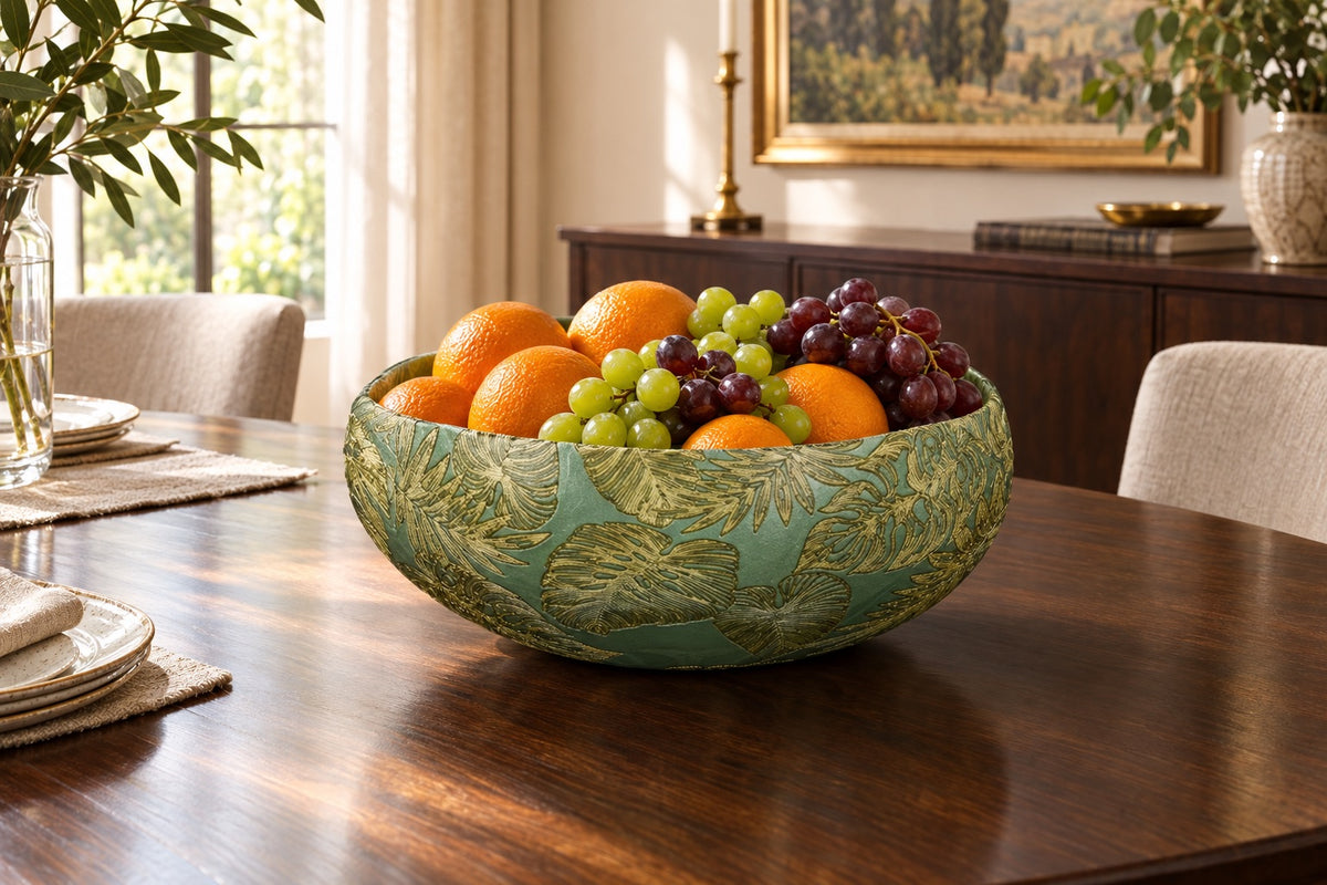 Grande coupe en verre verte à feuilles dorées remplie de fruits sur une table