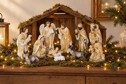 Ensemble de santons de crèche beige et or installés devant une maison de crèche en bois avec lumière dorée